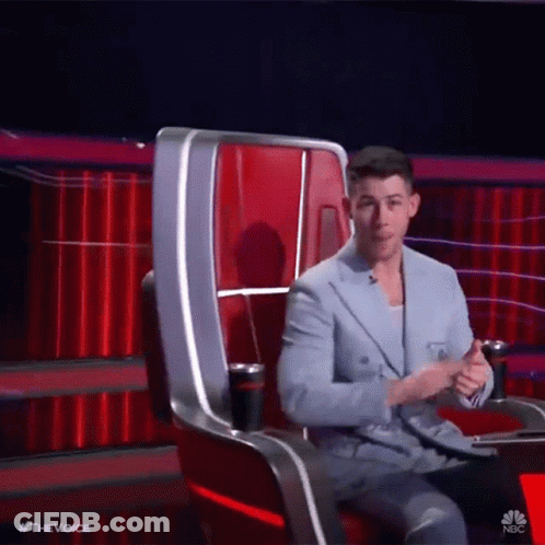 A Man In A Suit Is Sitting In A Chair On A Stage With A Red Curtain In The Background . GIF