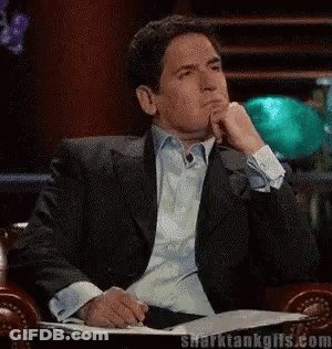 A Man In A Suit Is Sitting In A Chair With His Hand On His Chin And A Book In Front Of Him . GIF