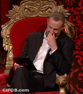 A Man In A Suit Is Sitting In A Red Chair Holding A Tablet And Covering His Mouth GIF