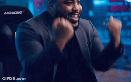 A Man In A Suit Is Sitting In An Akracing Chair With His Arms In The Air GIF