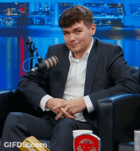 A Man In A Suit Is Sitting In Front Of A Microphone With A Supreme Bar On His Lap GIF
