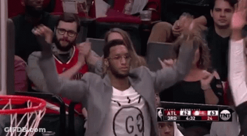 A Man In A Suit Is Sitting In The Stands At A Basketball Game With His Arms In The Air . GIF