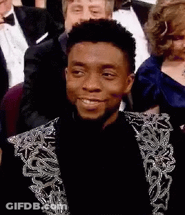 A Man In A Suit Is Smiling While Sitting In A Crowd . GIF