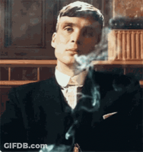 A Man In A Suit Is Smoking A Cigarette While Sitting At A Table . GIF