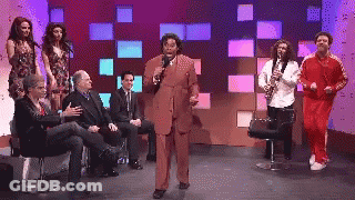 A Man In A Suit Is Speaking Into A Microphone While A Group Of People Sit Around Him GIF