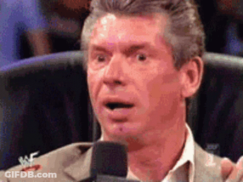 A Man In A Suit Is Speaking Into A Microphone With A Wrestler Logo On His Jacket GIF