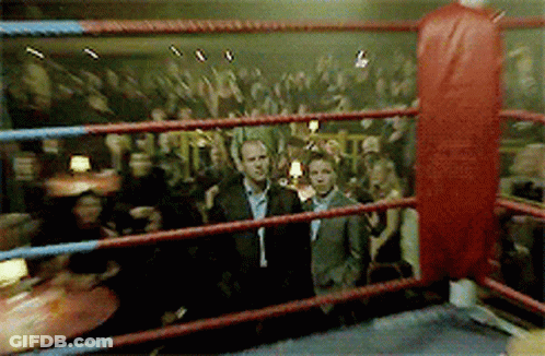 A Man In A Suit Is Standing In A Boxing Ring With A Crowd Behind Him GIF