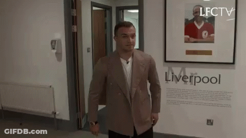 A Man In A Suit Is Standing In A Hallway Next To A Framed Picture Of A Man In A Red Shirt . GIF