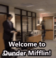 A Man In A Suit Is Standing In An Office With The Words Welcome To Dunder Mifflin Written On The Floor . GIF