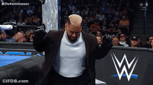 A Man In A Suit Is Standing In Front Of A WWE Sign GIF