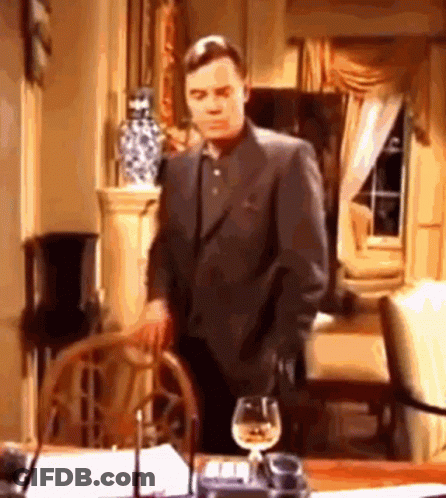A Man In A Suit Is Standing Next To A Table With A Glass Of Wine . GIF