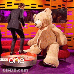 A Man In A Suit Is Standing Next To A Teddy Bear That Is Sitting On A Red Couch GIF