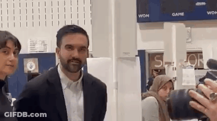 A Man In A Suit Is Standing Next To A Woman In A Hijab While Being Photographed By A Camera . GIF
