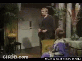 A Man In A Suit Is Standing Next To A Woman In A Purple Dress Who Is Sitting In A Chair GIF