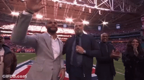 A Man In A Suit Is Standing On A Football Field With A Group Of People Applauding . GIF