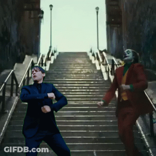 A Man In A Suit Is Standing On A Set Of Stairs Next To A Man In A Clown Suit GIF