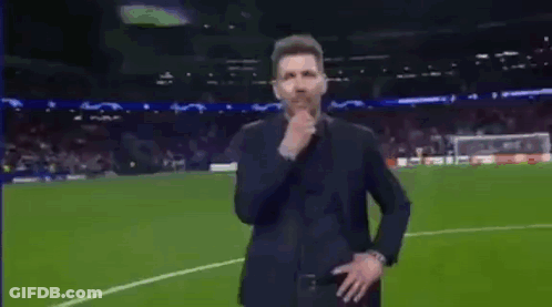A Man In A Suit Is Standing On A Soccer Field Holding His Hand To His Chin . GIF