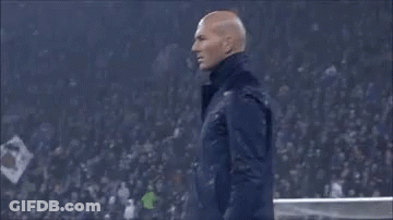 A Man In A Suit Is Standing On A Soccer Field Holding His Hand To His Head . GIF