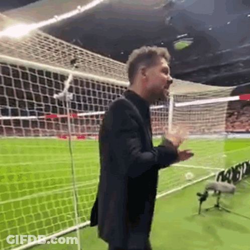 A Man In A Suit Is Standing On A Soccer Field With His Hands Outstretched . GIF