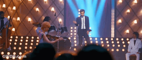 A Man In A Suit Is Standing On A Stage With A Woman Playing A Violin . GIF