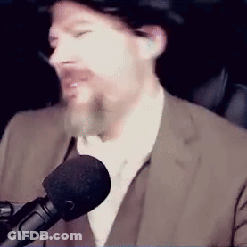 A Man In A Suit Is Talking Into A Microphone While Wearing A Hat . GIF