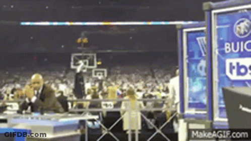 A Man In A Suit Is Talking On A Cell Phone In Front Of A Crowd At A Basketball Game Sponsored By Buick . GIF
