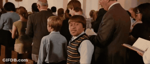 A Man In A Suit Is Talking To A Boy In A Striped Vest In A Church GIF