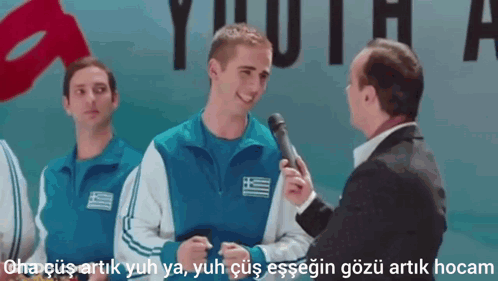 A Man In A Suit Is Talking To A Man In A Blue Jacket With A Greek Flag On His Chest GIF
