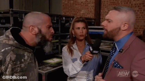 A Man In A Suit Is Talking To A Woman In A Microphone While Another Man Looks On . GIF