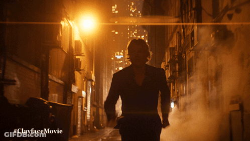 A Man In A Suit Is Walking Down A Dark Alleyway With The Hashtag #Clayfacemovie Below Him GIF