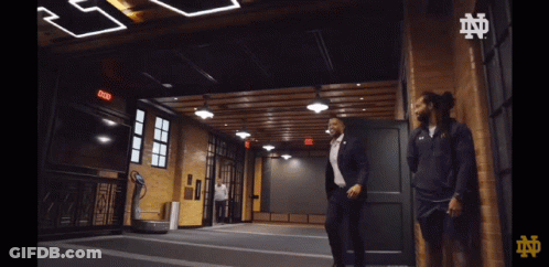 A Man In A Suit Is Walking In A Room With A Sign That Says Exit On It GIF