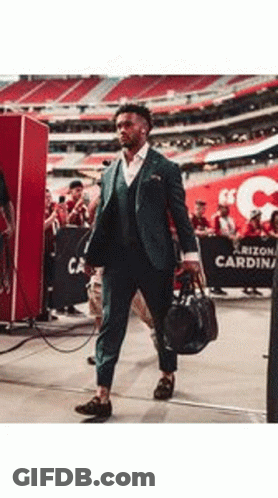 A Man In A Suit Is Walking Through A Stadium Holding A Bag . GIF