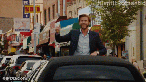 A Man In A Suit Is Waving From The Back Of A Car In A City GIF