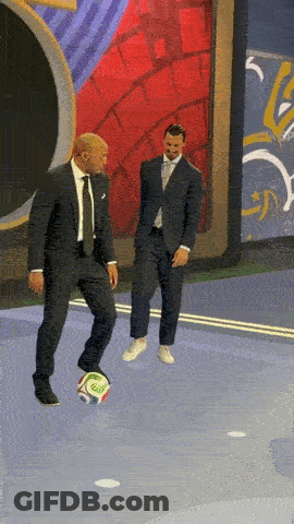 A Man In A Suit Kicks A Soccer Ball While Another Man Stands Behind Him GIF