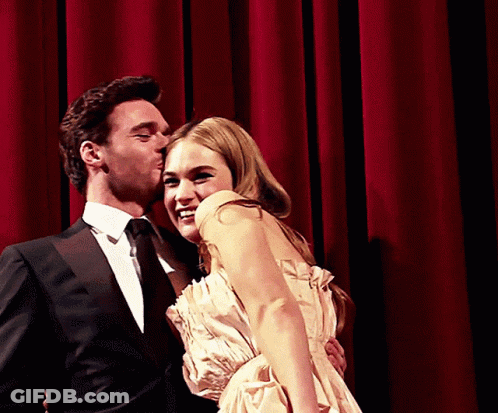 A Man In A Suit Kisses A Woman On The Cheek In Front Of A Red Curtain GIF