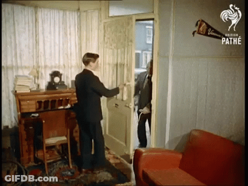 A Man In A Suit Opens A Door Next To A Red Couch GIF