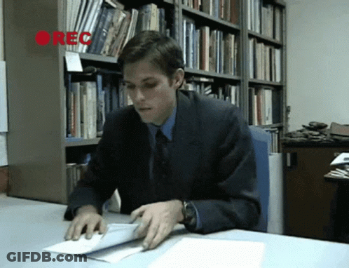 A Man In A Suit Sits At A Desk In Front Of A Bookshelf With The Word Rec On It GIF