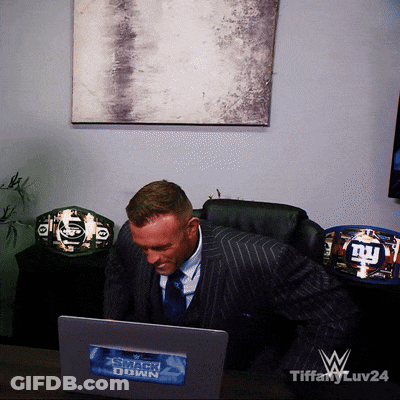 A Man In A Suit Sits At A Desk In Front Of A Laptop With A Sticker On It That Says Smackdown GIF