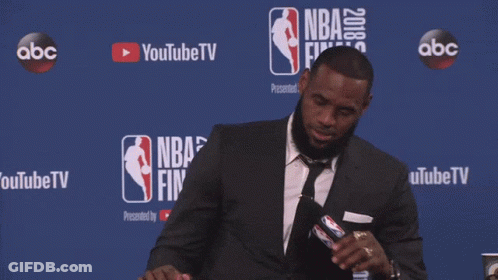 A Man In A Suit Sits At A Table In Front Of A Nba Finals Sign GIF