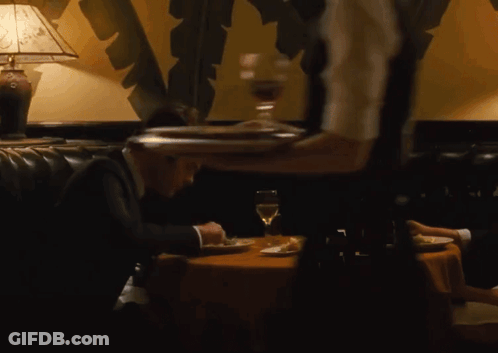 A Man In A Suit Sits At A Table While A Waiter Holds A Tray Of Food GIF
