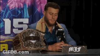 A Man In A Suit Sits At A Table With A Microphone And A Championship Belt On It GIF