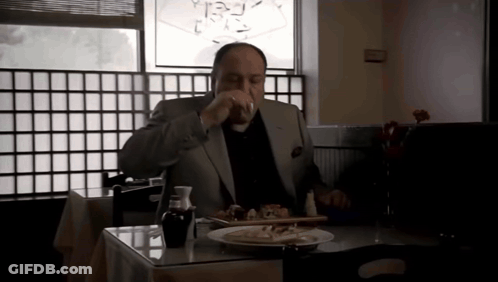 A Man In A Suit Sits At A Table With A Plate Of Food And A Glass Of Wine GIF