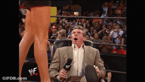 A Man In A Suit Sits In A Chair With A Microphone In Front Of A Wrestling Ring That Says Wwe On It GIF