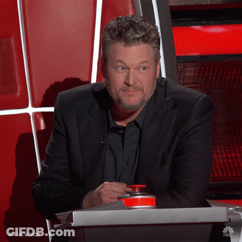 A Man In A Suit Sits In A Chair With A Red Button That Says The Voice On It GIF