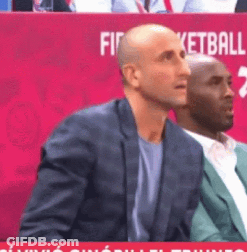 A Man In A Suit Sits In Front Of A Red Sign That Says Basketball GIF