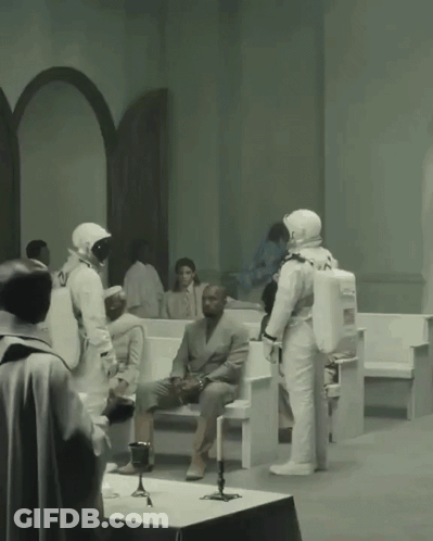 A Man In A Suit Sits On A Bench While A Group Of Astronauts Stand Around Him GIF
