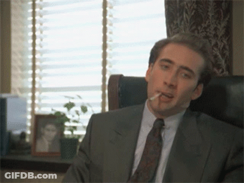 A Man In A Suit Smoking A Cigarette In Front Of A Window GIF