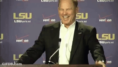 A Man In A Suit Stands At A Podium In Front Of A Wall That Says Lsu On It GIF