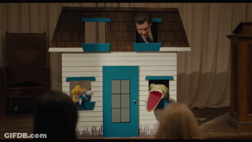 A Man In A Suit Stands Behind A Small House With A Puppet In The Window GIF