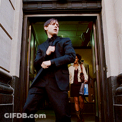 A Man In A Suit Stands In A Doorway While A Woman Walks In The Background GIF